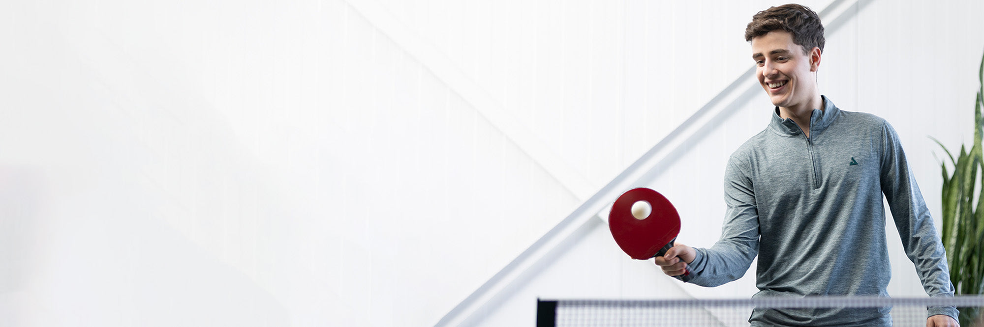 Junger Mann in grauem Langarmshirt spielt Tischtennis und schlägt Ball mit rotem Schläger.